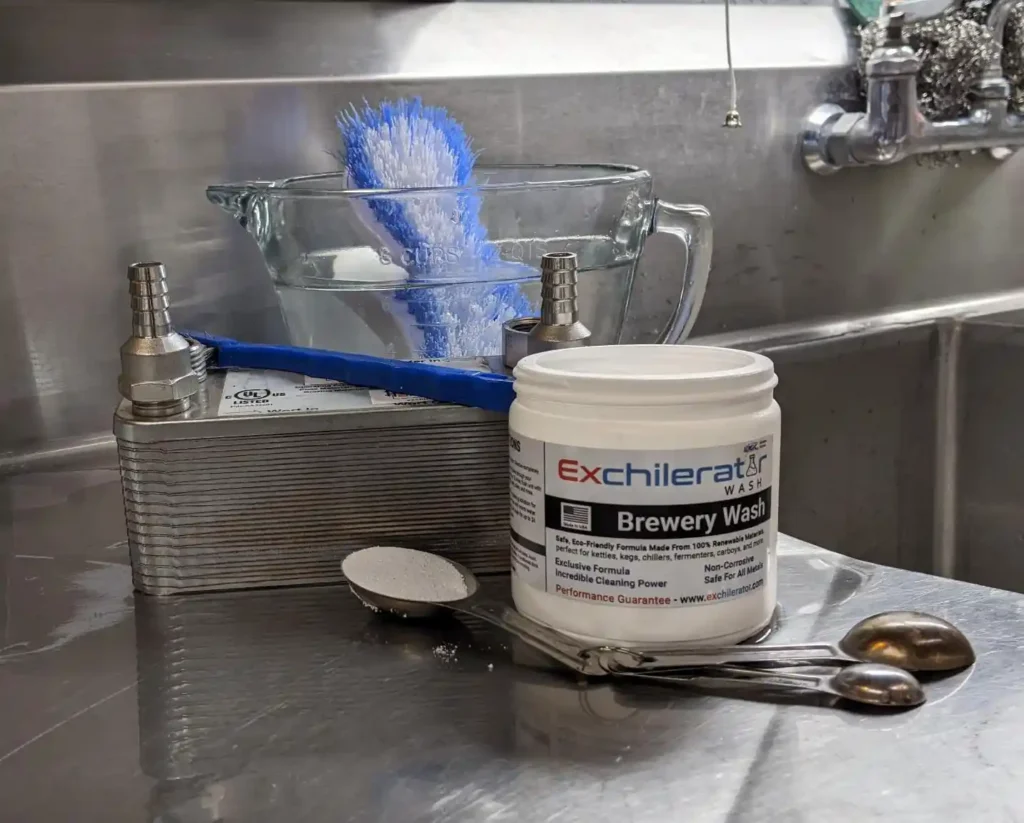 brewery wash, water, brush, and a plate chiller on a stainless steel sink getting ready to be cleaned.